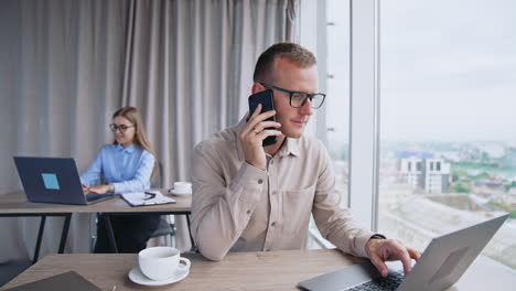 man holding phone at his ear waiting for answer. male employee puts off smartphone unsatisfied. female colleague prints on her laptop at backdrop.