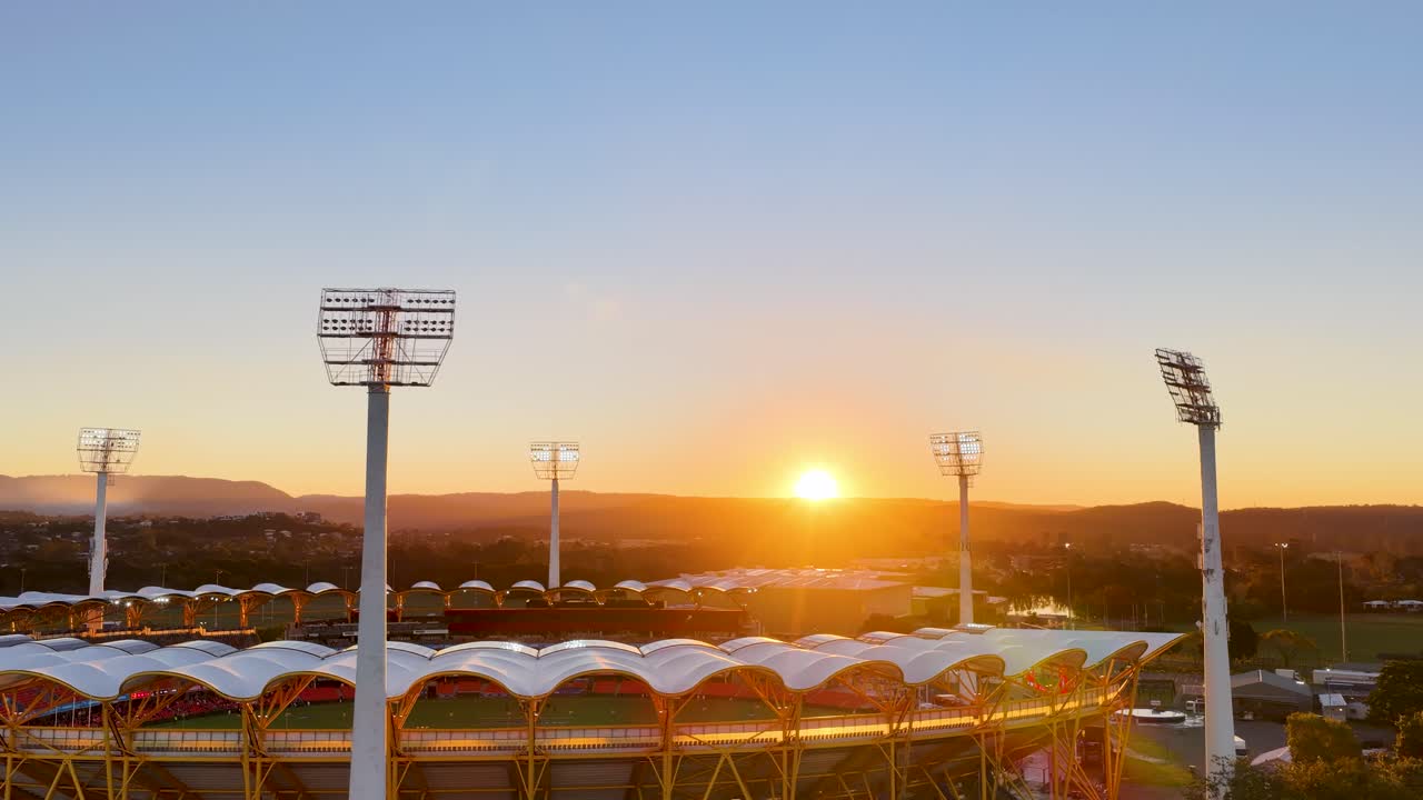 Drone captures a smooth, wide aerial view over a stadium at sunset in Gold Coast, Australia, with stadium lights and distant hills visible