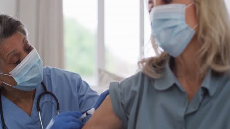 Senior-african-american-female-doctor-vaccinating-female-patient-at-home-both-wearing-face-masks