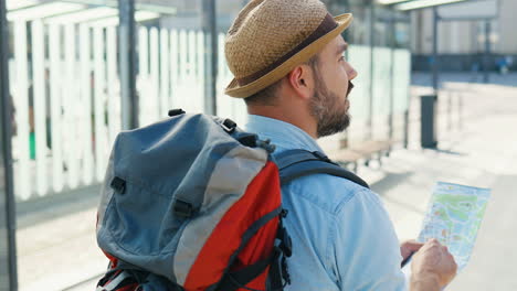 rear view of caucasian traveller wearing hat with backpack holding city map and walking down the street