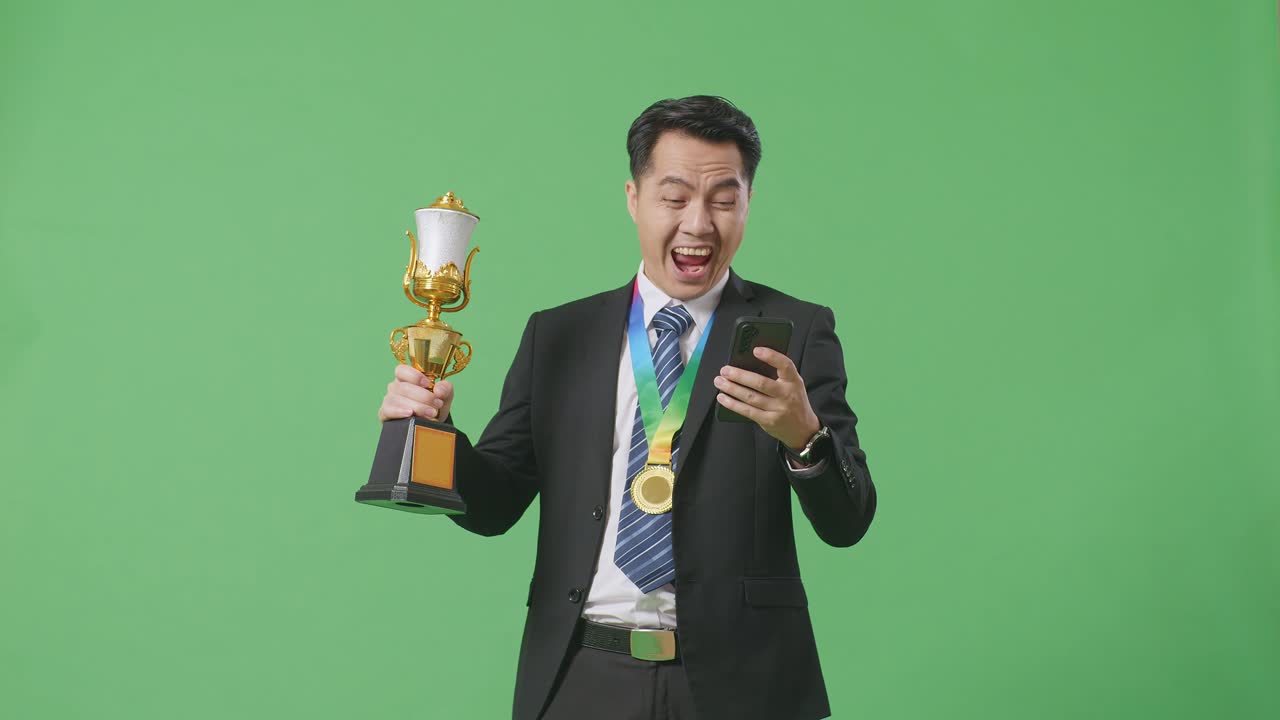 슈트와 타이를 입은 아시아 사업가, 금메달과 트로피를 들고 스마트폰을 쳐다보고, 그 다음에는 울부는 골, 스튜디오의 녹색 화면 배경에서 축하합니다.