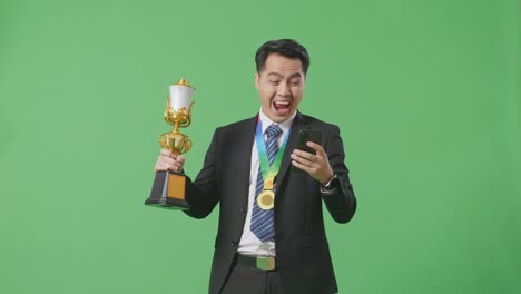 슈트와 타이를 입은 아시아 사업가, 금메달과 트로피를 들고 스마트폰을 쳐다보고, 그 다음에는 울부는 골, 스튜디오의 녹색 화면 배경에서 축하합니다.