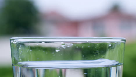 Wassertropfen-Treffen-Auf-Die-Oberfläche-Von-Kristallklarem-Wasser-In-Einem-Transparenten-Glasbecher-Auf-Einem-Verschwommenen-Hintergrund
