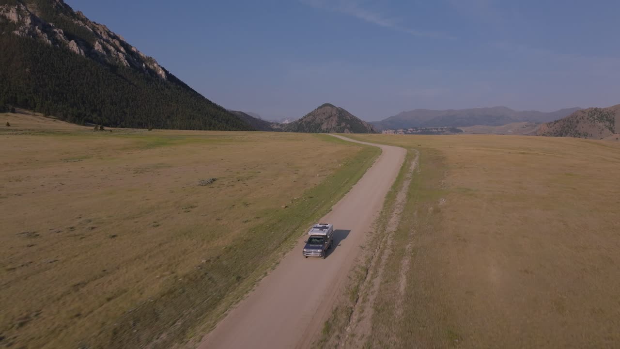 A lone vehicle drives on a dirt road through a vast open landscape under a clear blue sky