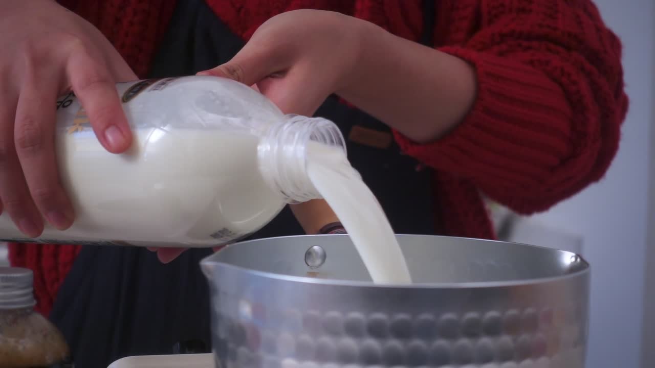 Close-up of milk being poured from a plastic bottle into a metal pot by hand, with warm indoor lighting and soft focus on texture and motion.