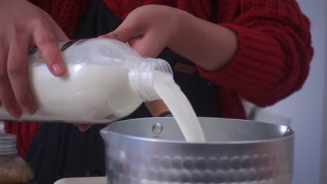 close-up of milk being poured from a plastic bottle into a metal pot by hand, with warm indoor lighting and soft focus on texture and motion.