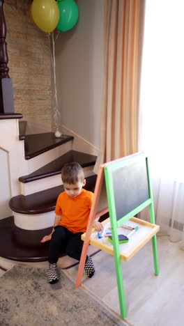 toddler boy and black cat sit on the stairs near the whiteboard. kid stands up and walks up to camera. high angle view. vertical video.