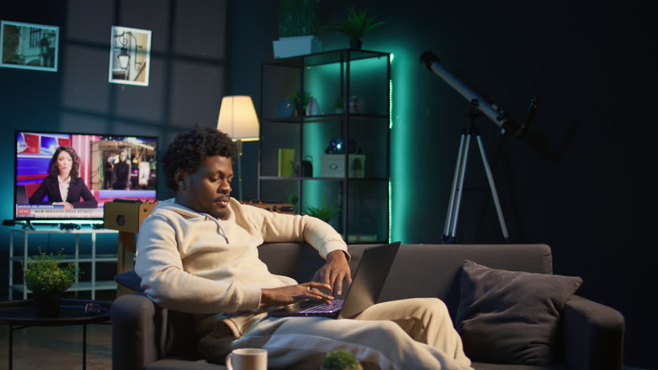 African American freelancer working remotely on a laptop under neon lights, sitting on a couch in a cozy loft apartment. Relaxed man in a stylish home highlights modern productivity. Camera B.