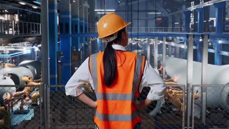 back view of asian female engineer with safety helmet in factory manufacture of wind turbines. standing with arms akimbo looking around, checking