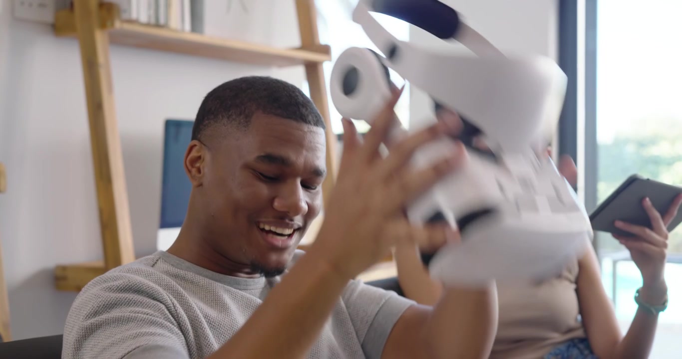 African American couple on couch at home, man with VR headset reaching, woman with tablet laughing