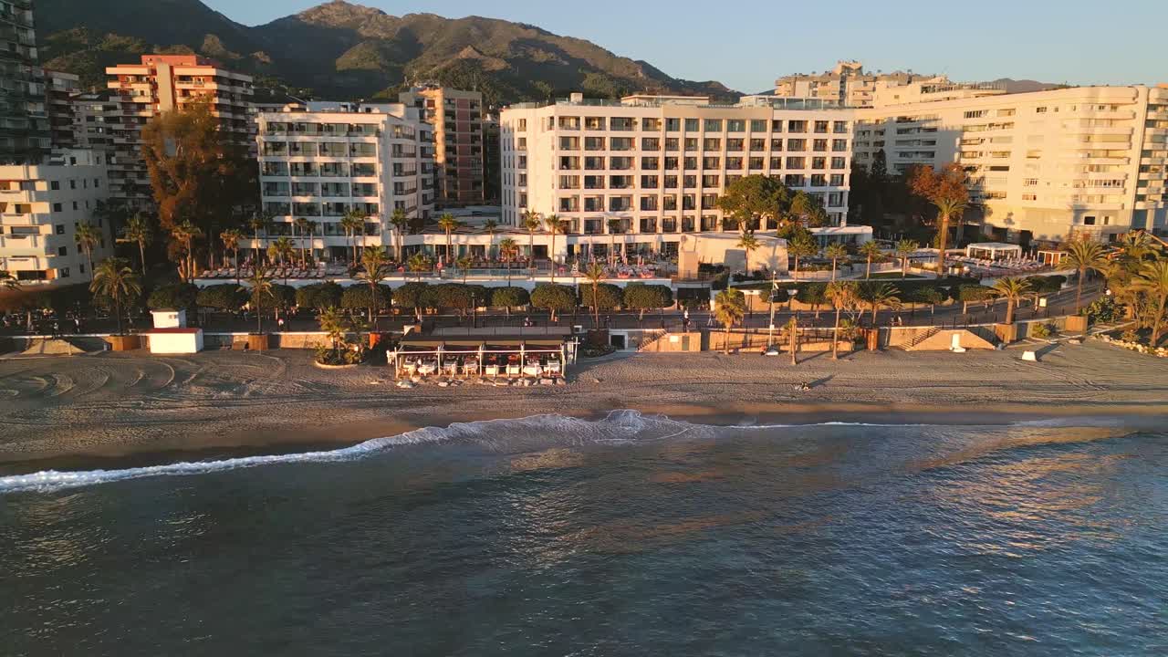 Ripresa aerea con carrello destro della meravigliosa spiaggia dorata di Marbella, del lungomare costeggiato da palme, degli eleganti edifici di lusso e di una vivace costa mediterranea al tramonto