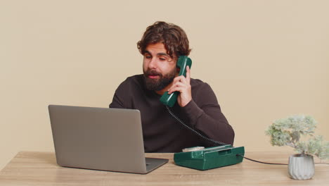 happy businessman sitting at table talking on wired telephone and using laptop on beige background