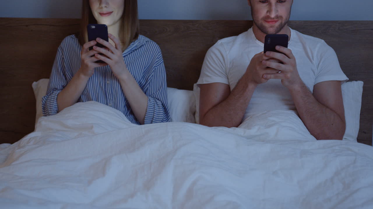 Caucasian couple using smartphones while they are sitting in the bed under the blanket