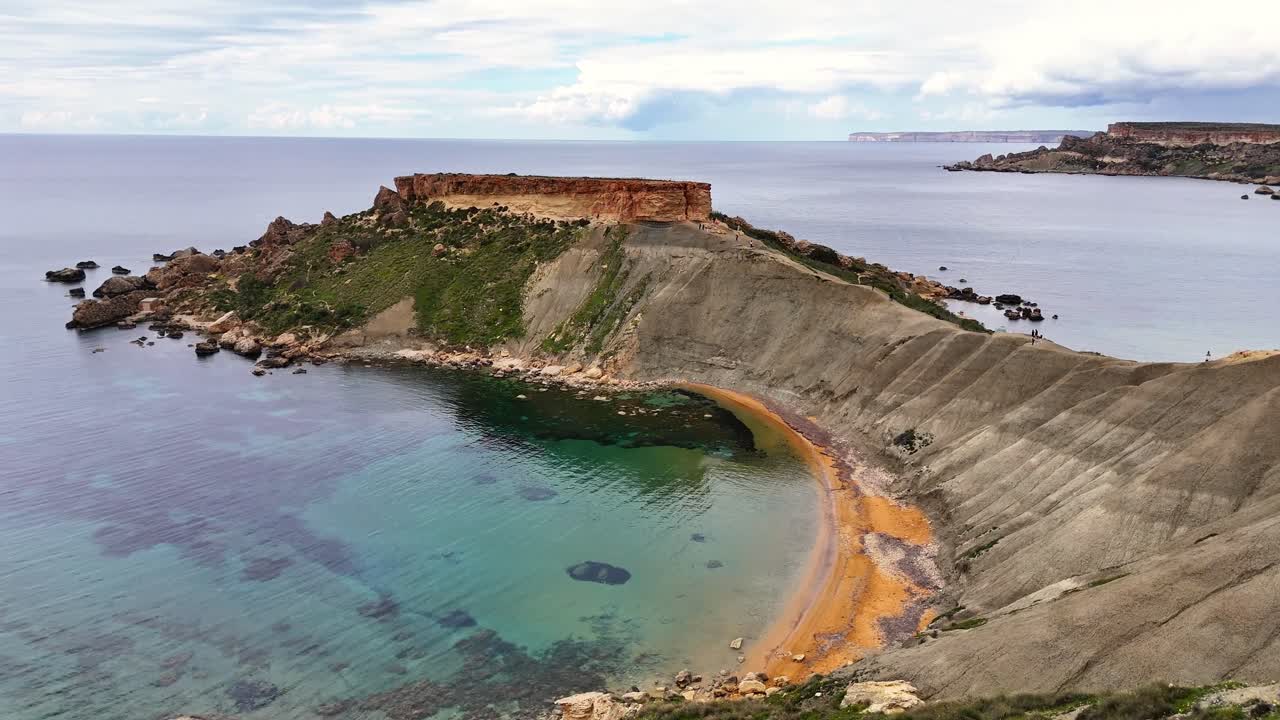 Vista aérea de falésias costeiras acidentadas e uma baía curva com águas claras na Baía de Qarraba, Malta, mostrando uma costa rochosa e terreno íngreme sob luz diurna suave.