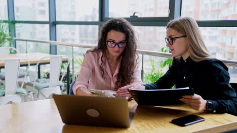 young female colleagues talking about working issues sitting at the table. ladies look at phone and have conversation.