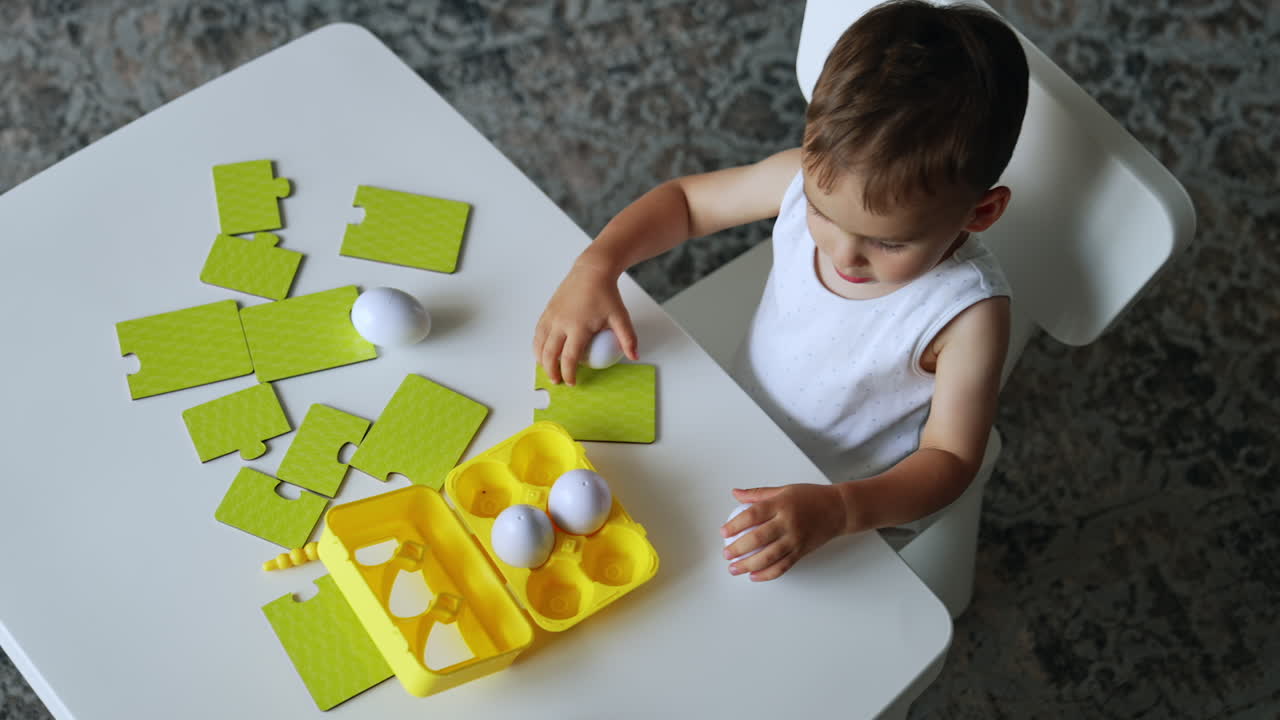 Caucasian toddler in a white t-shirt sits at desk. Kid plays with toy eggs and puzzles at once. Top view.