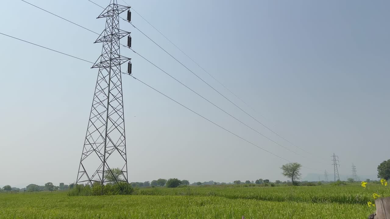 Tilt-up shot revealing a tall power transmission tower with long electrical lines stretching into a clear sky, farmland and distant trees grounding the scene below