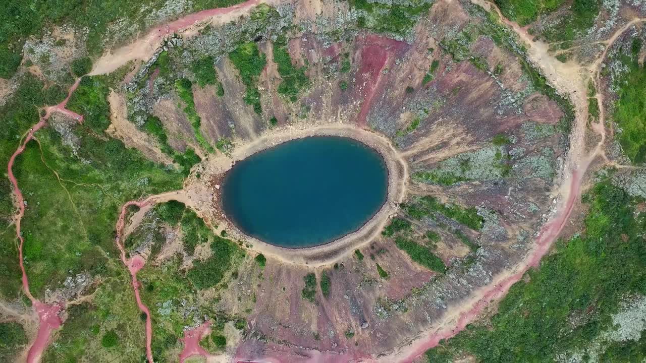 Aerial view of Kerið volcanic crater lake located in Grímsnes area in south Iceland, reflecting blue sky in the Golden Circle, featuring red slopes, green moss and hiking paths, drone top down view