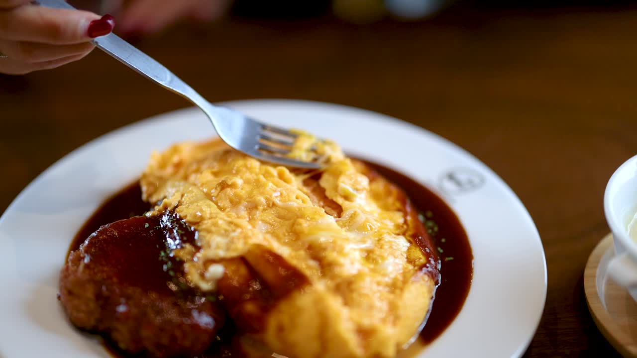 Hand slices cheesy Japanese omelette over hamburger steak with demi-glace, warm indoor lighting, close-up