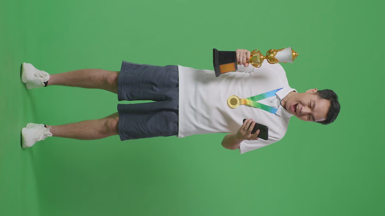 금메달과 트로피를 들고 스마트폰을 보고 있는 아시아 남자의 몸이 가득 차 있고, 그 다음에는 스튜디오의 녹색 스크린 배경에서 와우라고 말합니다.