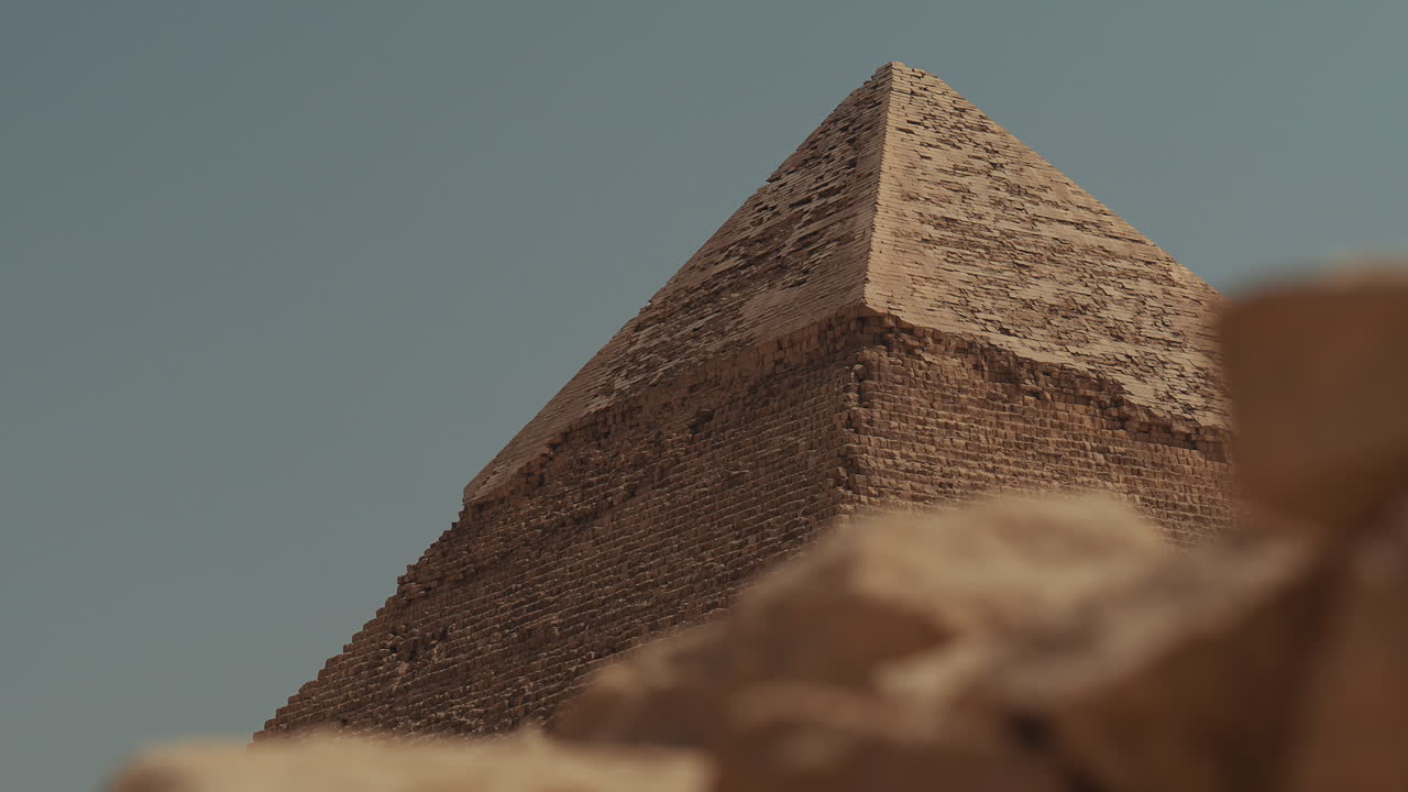 View of the Pyramid of Giza in Egypt with desert rocks in front and a clear blue sky above. The famous ancient wonder rises from the sandy landscape, showing the timeless history of this landmark