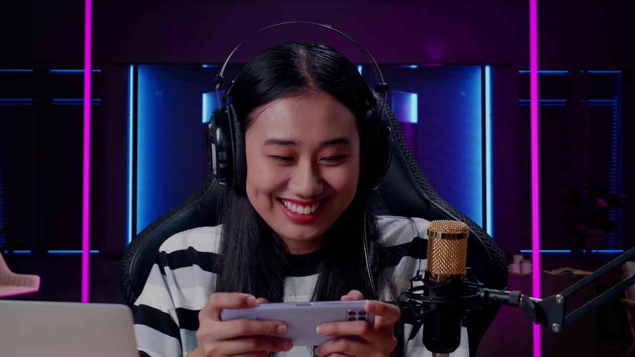 헤드폰과 컴퓨터를 가진 미소 짓는 아시아 여성 게이머가 휴대전화를 사용하여 레트로 네온 조명으로 조명된 방에서 게임을 하고 있습니다.