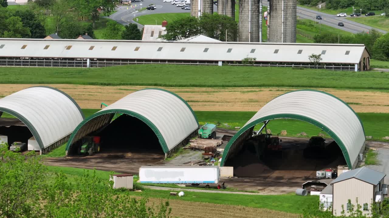 Aerial view of American agricultural structures with arched shelters and farm equipment. Lush fields illustrate vibrant rural landscape, showing modern U.S. farming operations. Barn with silo storage