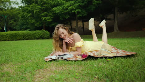 Feliz-Mujer-Sonriente-Vestida-De-Amarillo-Disfruta-Leyendo-Un-Libro-Tumbado-En-El-Césped-Verde-En-Un-Picnic-En-El-Parque-De-Verano