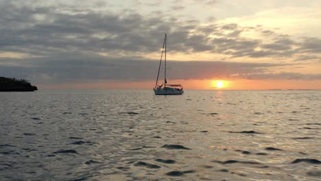 Vista-Aérea-Cinematográfica,-Velero-Anclado-En-El-Mar-Por-La-Costa-De-La-Isla-Con-Una-Pintoresca-Puesta-De-Sol-En-El-Horizonte,-Disparo-De-Drones