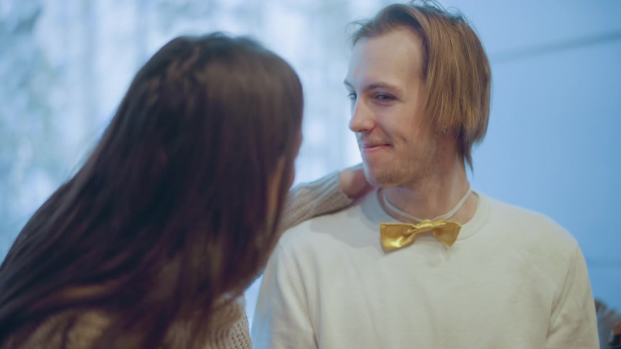 Close up of woman adjusting man gold bow tie while standing indoors with soft lighting. Man wears white shirt scene features calm interaction and clear focus on accessory adjustment