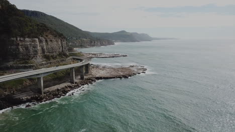 Antena:-Dron-Descendiendo-Mientras-Un-Automóvil-Atraviesa-Un-Puente-De-Carretera-Junto-Al-Océano-En-Nueva-Gales-Del-Sur,-Australia