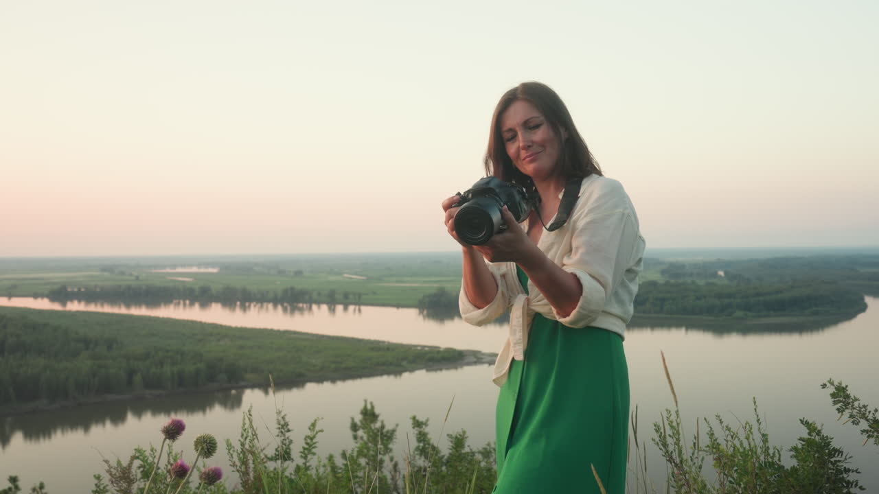 woman dressed in white shirt and green skirt smiles while taking photo with camera in hands, standing in grassy field overlooking calm river and distant forest
