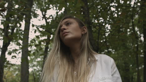 Low-angle-still-shot-young-blonde-woman-looking-up-at-trees-in-nature-forest-environment