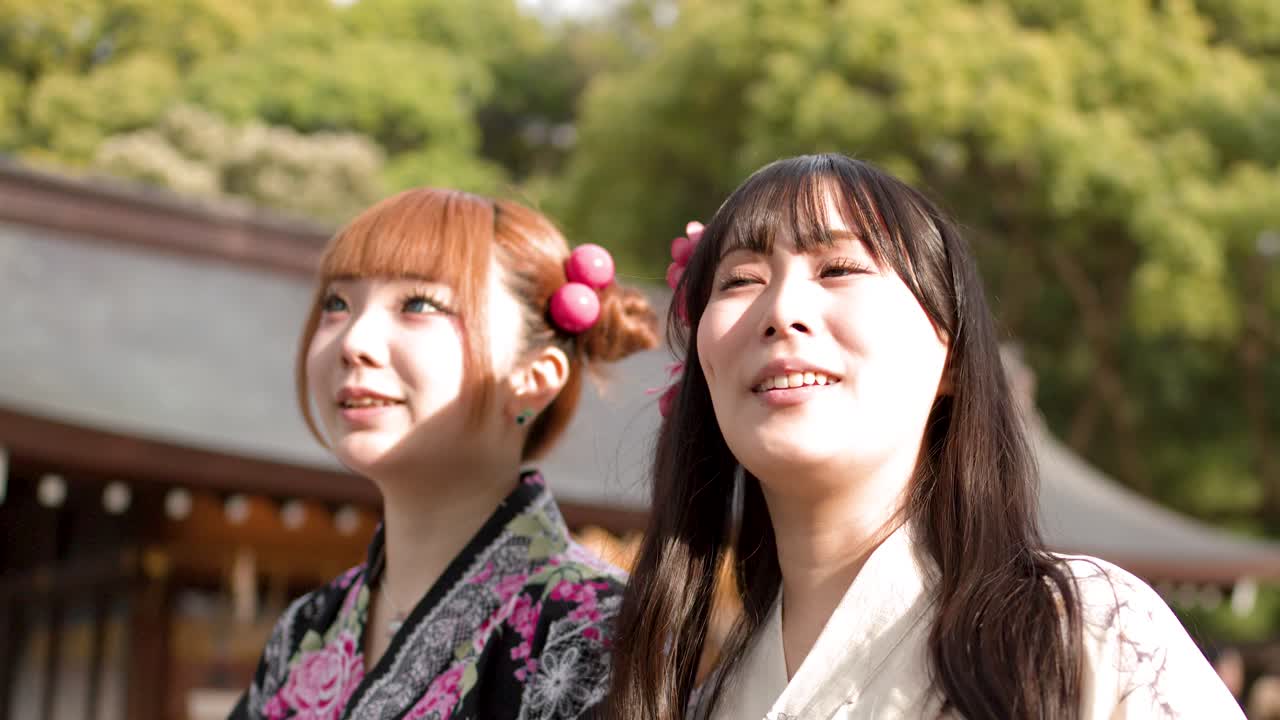 Duas mulheres japonesas usando yukata tradicional sorriem e conversam ao ar livre em um santuário xintoísta.