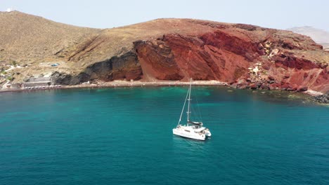 antena descendiendo sobre la playa roja con velero pasando por santorini, grecia