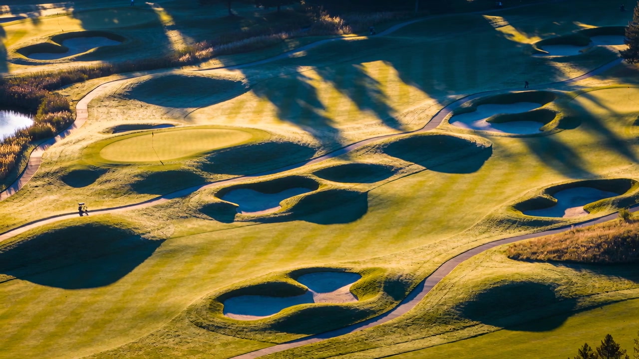 Toont gevormde potbunkers onder lage zon, golfers lopen over het karrenpad richting de green op de golfbaan