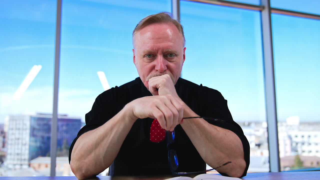 Adult Caucasian businessman wearing black clothes and red tie sits at desk. Positive man working in spacious office. Low angle view.