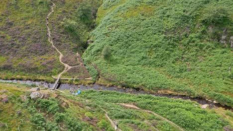 aerial drone video footage of a footpath and stream, river showing a small bridge structure with people resting along the riverbank. shot in near the village if marsden in the united kingdom