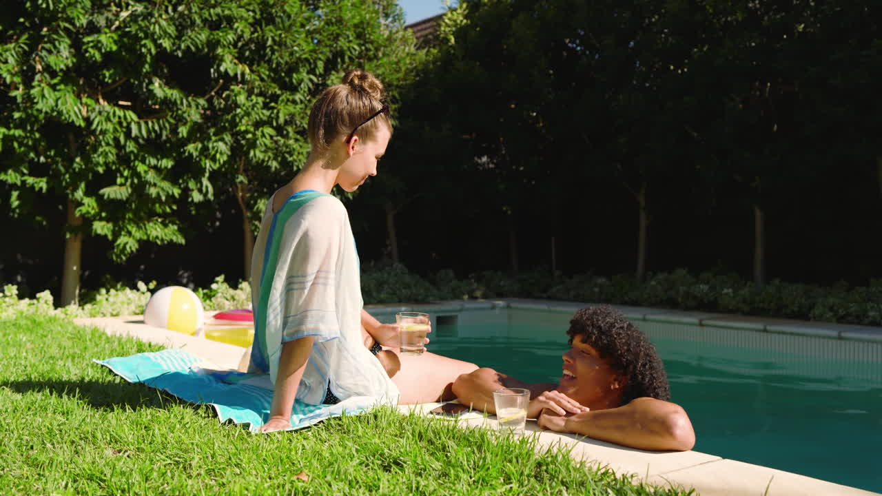 Relaxing by pool, diverse couple enjoying drinks and sunshine, sharing conversation, copy space