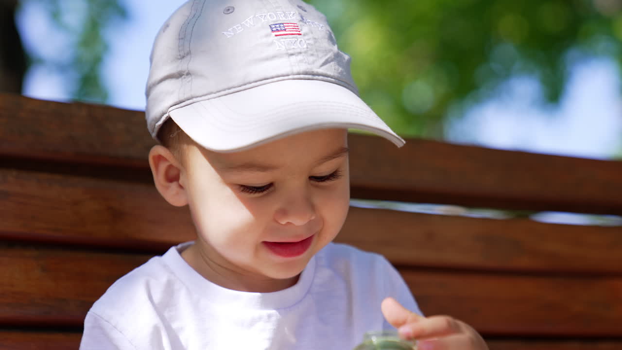 Beautiful Caucasian toddler in cap with a water bottle in hands. Thirsty child on the bench outdoors in summer. Close up.