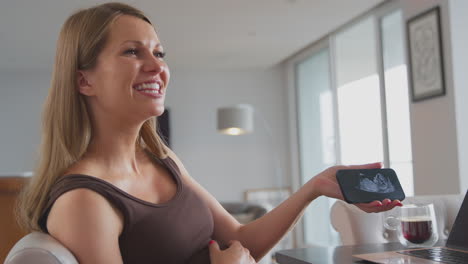 smiling pregnant woman at home holding mobile phone with ultrasound scan of baby