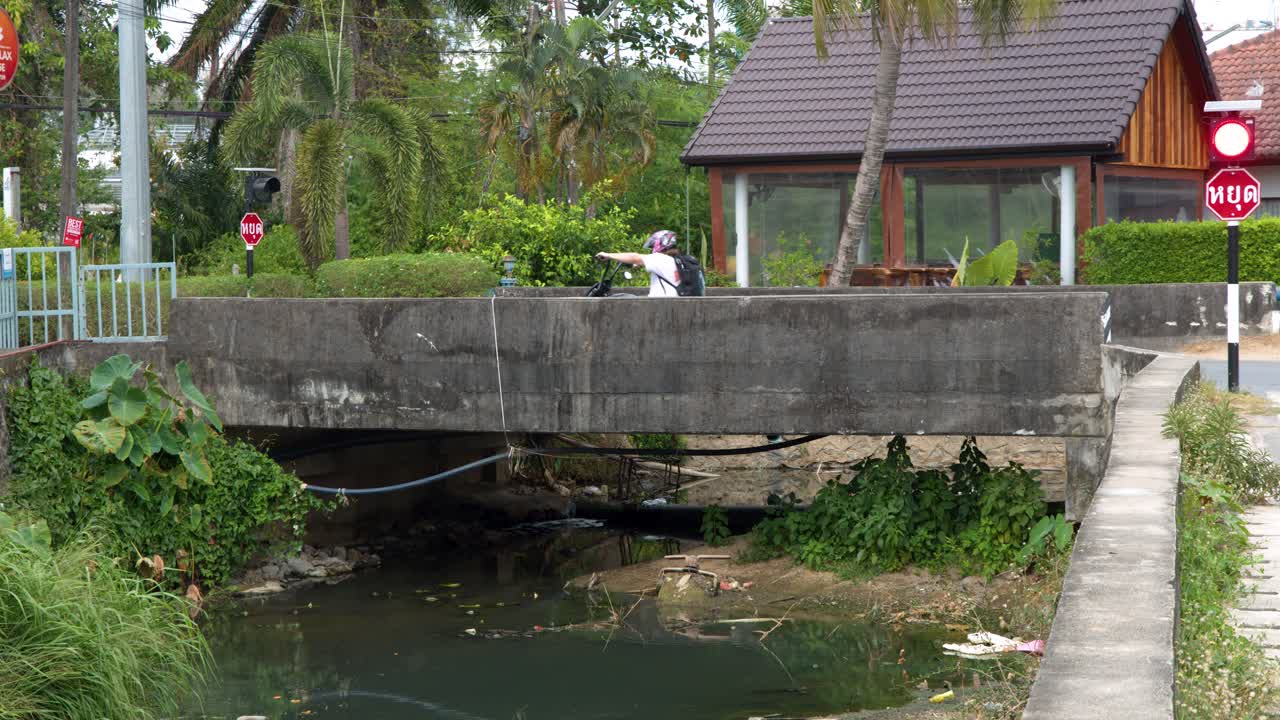 En vidvinkelbild visar en motorcyklist som korsar en betongbro över en förorenad thailändsk kanal.