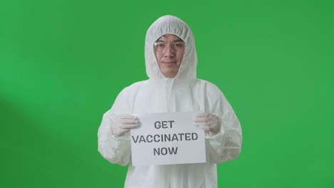 asian man wearing personal protective equipment uniform ppe and holding get vaccinated now sign in the green screen studio
