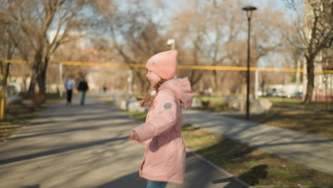 little girl in a pink cap and jacket, wearing blue jeans, joyfully chases pigeons on a sunny park pathway