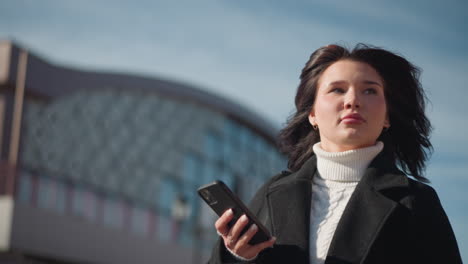 confident woman outdoors in black coat and white turtleneck holding smartphone, looking around with determination against modern building background focus back on her phone