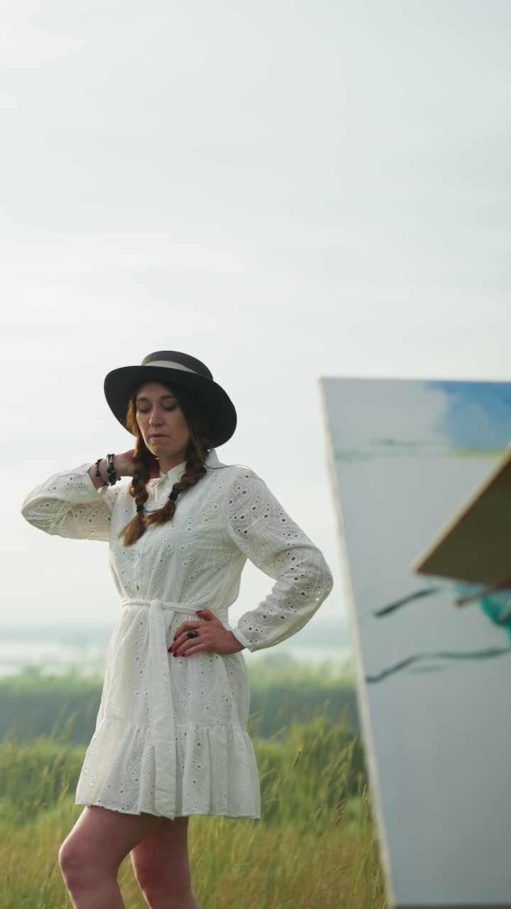 A woman in a white dress and hat poses elegantly in a grassy field while an artist in a checkered shirt and jeans, holding a palette, paints her portrait
