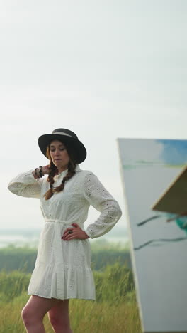 a woman in a white dress and hat poses elegantly in a grassy field while an artist in a checkered shirt and jeans, holding a palette, paints her portrait