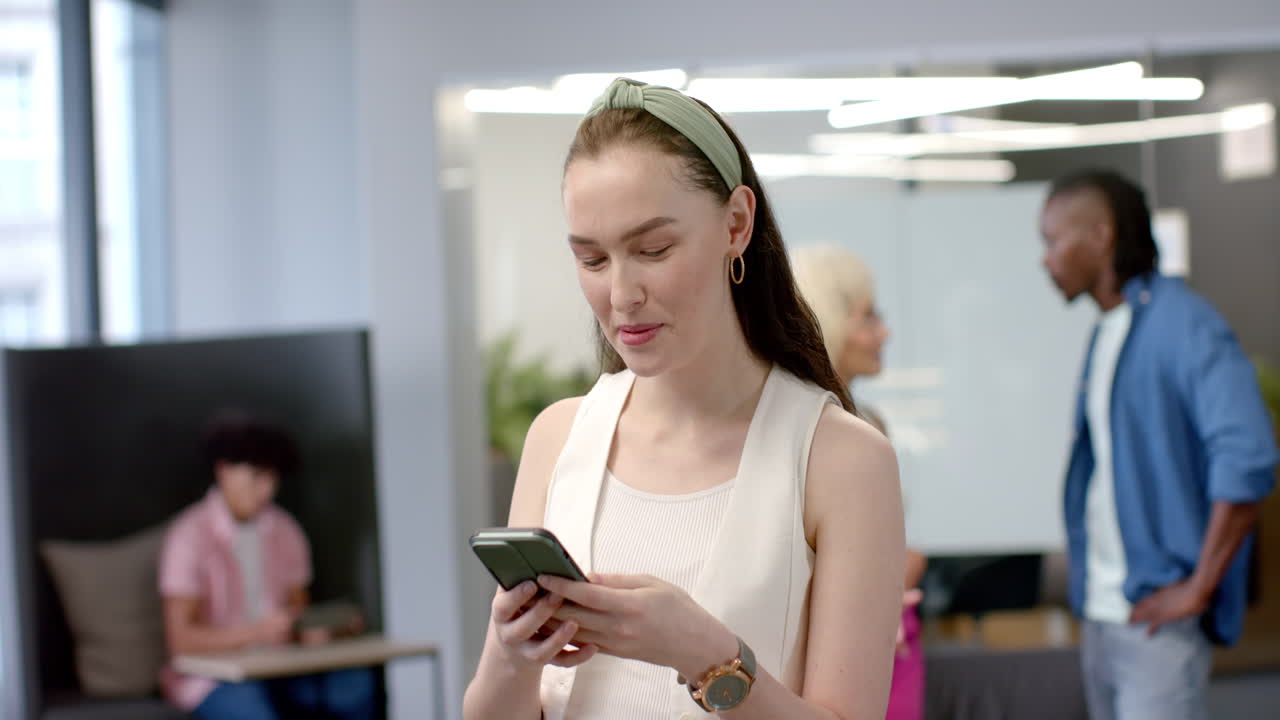 Using smartphone, woman standing in office with colleagues in background