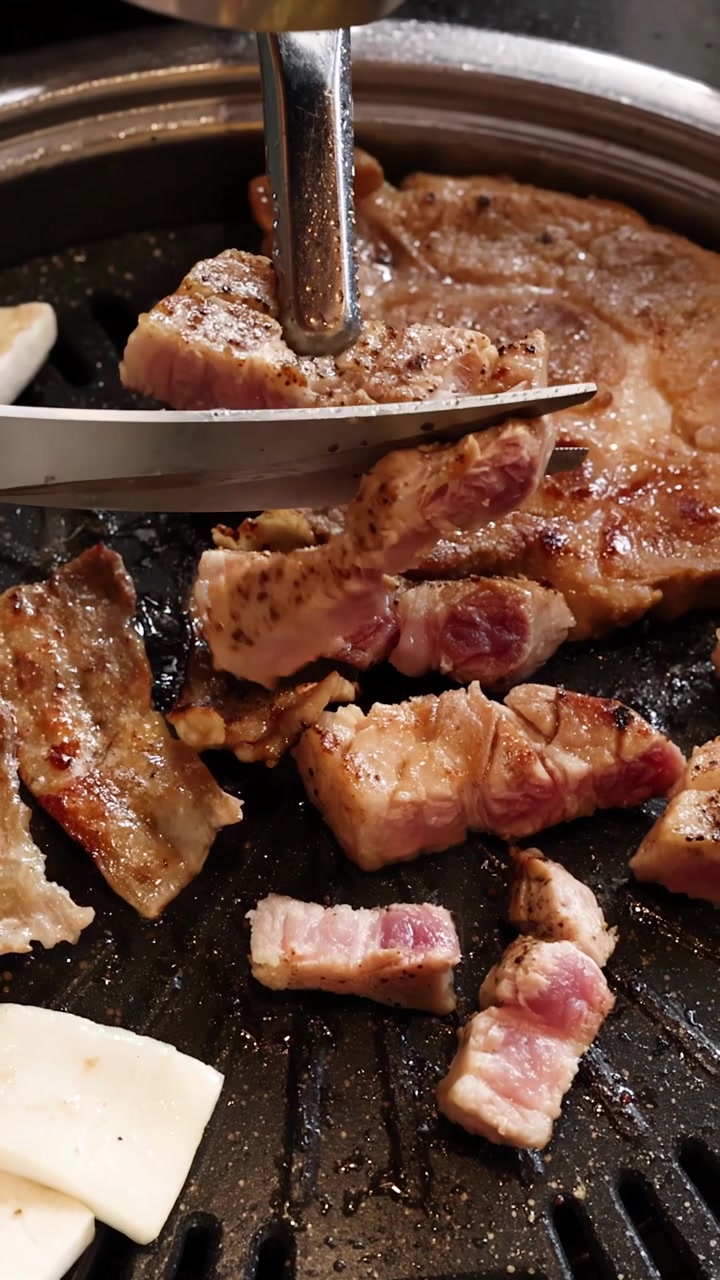 Tangan menggunakan penjepit dan gunting untuk memotong samcan babi di atas panggangan barbekyu Korea panas, close-up