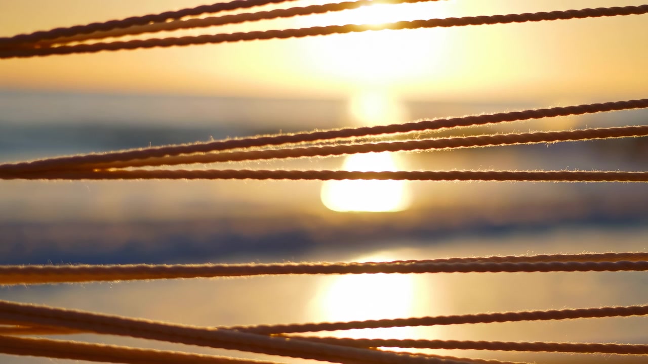 La calda luce serale del sole brilla sull'acqua mentre cavi paralleli attraversano l'inquadratura, mostrando tenui riflessi, foschia all'orizzonte lontano e una vista costiera pacifica con colori tenui e illuminazione delicata.